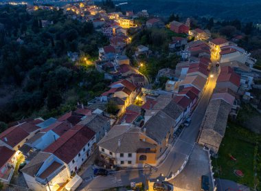 Geceleri Korfu Yunanistan 'da Agrafoi köy meydanı ve sokaklarının panoramik hava manzarasında geleneksel binalar ve alacakaranlıkta sokak lambaları yer alıyor.