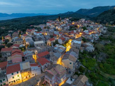 Yunanistan 'ın Korfu adasındaki dağlarda geleneksel mimari ve dolambaçlı yolların yer aldığı, aydınlanmış Nimfes köyünün alacakaranlıktaki hava manzarası
