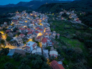 Aerial view of illuminated Nimfes village at dusk, featuring traditional architecture and winding roads in the mountains of Corfu island, Greece