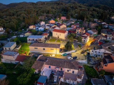 Panoramic drone shot of Valanio traditional settlement in Corfu island Greece. Scenic mountain landscape with illuminated village homes, cypress trees and lush forest at twilight evening.