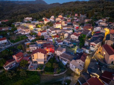 Yunanistan Korfu adasında geleneksel Valanio yerleşiminin panoramik insansız hava aracı çekimi. Aydınlatılmış köy evleri, selvi ağaçları ve alacakaranlık vakti yemyeşil ormanlarıyla manzaralı bir dağ manzarası..