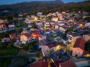 Yunanistan Korfu adasında geleneksel Valanio yerleşiminin panoramik insansız hava aracı çekimi. Aydınlatılmış köy evleri, selvi ağaçları ve alacakaranlık vakti yemyeşil ormanlarıyla manzaralı bir dağ manzarası..