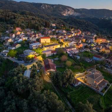 Korfu adasındaki Valanio köyünün hava aracı manzarası alacakaranlıkta aydınlatılmış sokaklar ve yeşil dağlara kurulmuş geleneksel evlerle Yunanistan