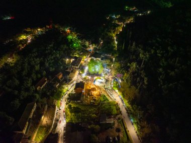 Nymfes, Corfu, Yunanistan 'da düzenlenen geleneksel köy festivalinin gece görüşünde geniş bir ışıklandırılmış ağaç, kalabalık bir meydan, canlı bir kutlama ve yol boyunca park edilmiş arabalar yer alıyor..