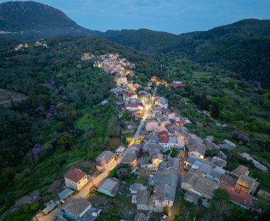 Panoramic aerial drone view of Zigos village in Corfu Greece at twilight featuring illuminated traditional houses and a vast green mountain landscape