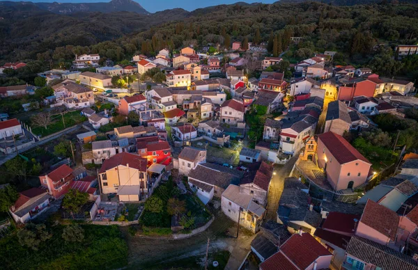 Yunanistan Korfu adasında geleneksel Valanio yerleşiminin panoramik insansız hava aracı çekimi. Aydınlatılmış köy evleri, selvi ağaçları ve alacakaranlık vakti yemyeşil ormanlarıyla manzaralı bir dağ manzarası..