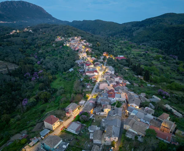 Panoramic aerial drone view of Zigos village in Corfu Greece at twilight featuring illuminated traditional houses and a vast green mountain landscape