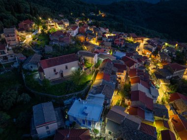 Episkepsi, Korfu, Yunanistan 'daki dağ yerleşiminde gece aydınlatılan geleneksel bir ortodoks kilise ve köy meydanının tepeden aşağı insansız hava aracı görüntüsü