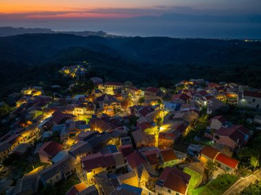 Yunanistan 'ın geleneksel dağ köyü Episkepsi' nin insansız hava aracı görüntüsü alacakaranlıkta parlayan sokak ışıkları ve renkli günbatımı gradyan gökyüzü, Korfu, Yunanistan