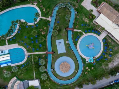 Güneşli bir yaz tatili sırasında Yunanistan 'da bir tatil beldesinde yeşil çimenler ve yüzme havuzları arasında kıvrılan tembel bir nehri olan bir su parkının tepeden aşağı insansız hava aracı görüntüsü