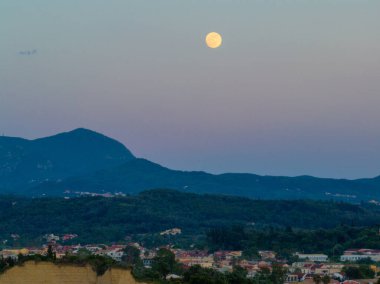 Korfu 'daki uzak dağ sırtları ve banliyö manzarasının üzerinde parlayan dolunayın atmosferik akşam görüntüsü.