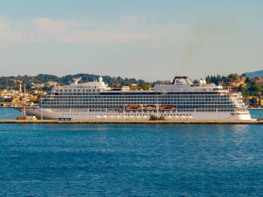 Korfu Yunanistan rıhtımına demirlemiş beyaz Viking Denizi yolcu gemisinin tam yan görüntüsü, geminin uzunluğunu ve balkonlarını ağaçlar ve geleneksel binalara karşı sergiliyor.