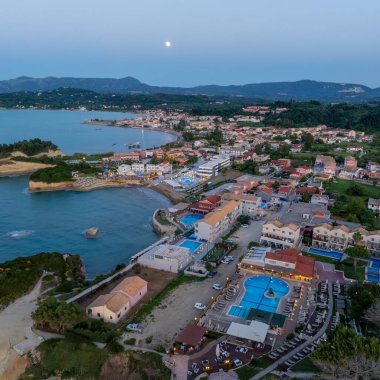 Korfu Yunanistan 'daki Sidari sahil beldesinin hava manzarası alacakaranlıkta aydınlık oteller, yüzme havuzları ve İyon Denizi kayalıklarının üzerinde yükselen ay ile birlikte..