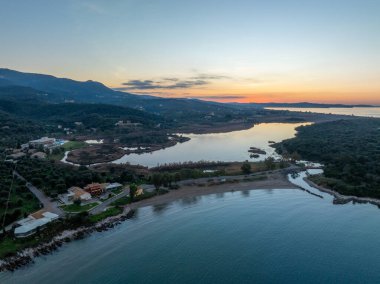 Agios Spiridon plajı üzerindeki canlı günbatımı manzarası ve Korfu Yunanistan 'daki Antinioti gölünde ufukta deniz ve dağlarla buluşan kumlu bir kıyı şeridi yer alıyor.