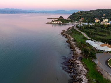 Alacakaranlıkta sakin deniz suyuyla birlikte yumuşak pembe pastel gökyüzü altında Agios Spiridon North Corfu Yunanistan 'daki kayalık kıyı şeridi ve kıyı yolunun manzaralı manzarası