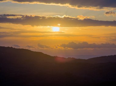Yunan adalarındaki siluetli bir dağ sırasının üzerinde mercek parıltısı ve altın bulutlarla parlak bir günbatımı gökyüzü.