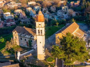 Yunanistan 'ın Korfu Adası' ndaki Potamos köyünde yüksek çan kulesi ve mezarlığı olan geleneksel bir Ortodoks kilisesinin havadan görünüşü, sıcak günbatımı ışığı ve yeşil çevresiyle altın saat içinde çekildi.