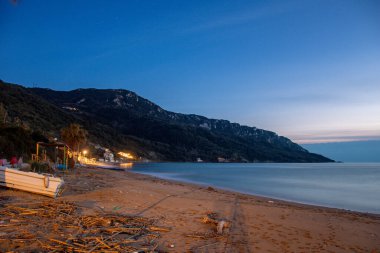 Mavi saat boyunca Korfu Yunanistan 'da kumlu bir sahilde dinlenen küçük beyaz bir tekne. Sakin deniz, kıyı dağları ve gece ışıkları su yüzeyine yansıyor.