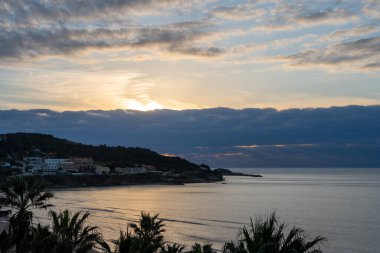 Korfu 'nun Agios Stefanos plajında çarpıcı bir gündoğumu manzarası var. Yunanistan' da siluetli palmiye ağaçları, ağır bulutlar, arka planda deniz ve kıyı köylerindeki evleri yansıtan altın güneş ışığı bulunuyor.