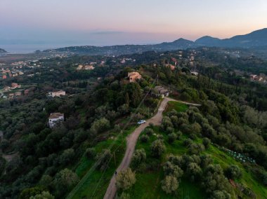 Korfu 'da günbatımında evlerin ve zeytin ağaçlarının bulunduğu bir tepenin kenarındaki dolambaçlı bir yolun hava manzarası.