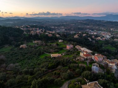 Korfu 'nun yemyeşil geleneksel bir köyünün hava panoramik manzarası uzak dağlarla dolu renkli bir günbatımı sırasında Yunanistan zeytin korulukları ve tepelerin arasına yuva yapmış durumda