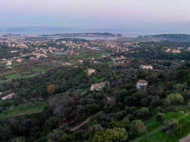 Korfu 'daki bir kıyı kasabasının panoramik hava aracı görüntüleri, Yunanistan' ın yeşil tepeler arasında dağınık evleri ve yumuşak bir akşam gökyüzünün altındaki uzak denizi gösteriyor.