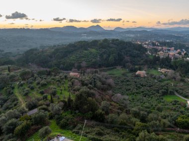 Güneş batarken Korfu kırsalının manzaralı manzarası. Yuvarlanan tepeler, yoğun zeytin ormanları ve sıcak turuncu ve mavi alacakaranlık gökyüzü altında dağınık evler.