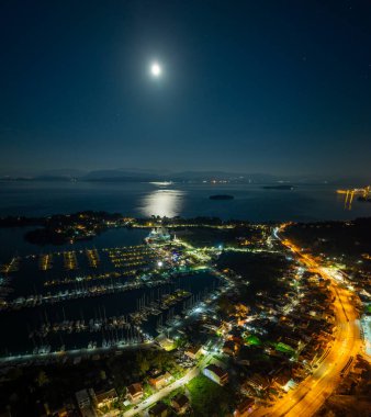 Yunanistan 'ın Korfu Adası' ndaki Gouvia Limanı 'nın hava panoramik manzarası. Dolunay İyon Denizi' ni ve şehir ışıklarını yansıtıyor. Sahil şeridini, limanı ve demirlemiş yatları aydınlatıyor.