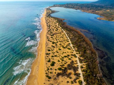 Halikounas plajının kum tepelerinden geçen toprak yolun yüksek açılı hava aracı görüntüsü, mavi denizi Yunanistan 'ın Korfu kentindeki Korission Gölü' nün sakin sularından ayırıyor..