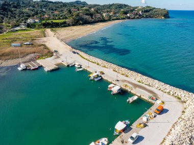 Hava aracı Corfu adasındaki küçük geleneksel balıkçı limanının tepesinde turkuvaz suyuna bulanmış renkli tekneler, taş kırıcı su koruması, Yunan kıyı köyü manzarası.