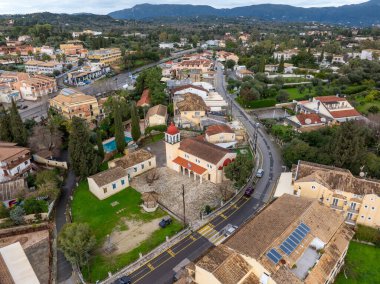 Gouvia Korfu Yunanistan 'daki Agion Panton Ortodoks Kilisesi' nin hava görüntüsünde kırmızı bir kubbe, çan kulesi, taş avlu ve üzerinde boyalı taksi işaretleri olan bir cadde var.