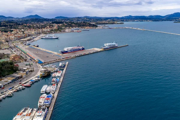Yunanistan 'ın Kerkyra kentindeki Korfu Yeni Limanı' nın hava manzarasında yolcu feribot gemileri, seyir terminalleri ve bulutlu bir gökyüzü altında dağ ve kentsel mimari manzaralı İyon Denizi kıyı şeridi yer alıyor..