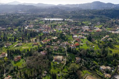 Korfu 'nun geleneksel kırsal bir köyünün panoramik hava aracı görüntüsü. Dağılmış evler, yemyeşil tepeler ve arka planda gölleri ve dağları olan zeytinlikler arasında.