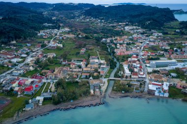 Yunanistan 'ın Korfu kentindeki Sidari kıyı köyünün havadan panoramik manzarası, İyon Denizi, konut evleri, oteller ve bulutlu bir kış gökyüzü altında yemyeşil tepelerin buluştuğu nehri gösteriyor..