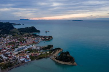 Sidari, Korfu, Yunanistan 'daki Kanal d' Amour 'un hava görüntüsü, sahil kenti ile günbatımında eşsiz kum taşı kaya oluşumları ve turkuaz İyon Denizi' ni gösteriyor..