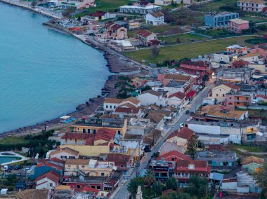 Yunanistan 'ın Korfu kentinde canlı bir sahil kasabasının hava aracı perspektifi, renkli binalar, geleneksel kırmızı kiremitli çatılar, kıyı yolları ve İyon Denizi kıyısında kayalık bir kıyı şeridi yer alıyor..
