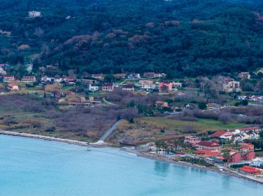 Yunanistan 'ın Corfu kentindeki sakin İyon Denizi kıyı şeridi boyunca yoğun yeşil zeytin koruluklarına ve tepelere yuva yapmış sakin bir kıyı köyünün havadan görünüşü.