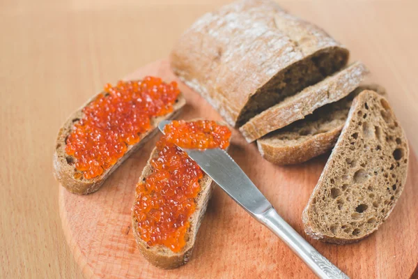 Red caviar toasts sandwich with silver knife, bowl of tasty red caviar with spoon and fresh baked homemade healthy bread on a wooden table background — Stock Photo, Image