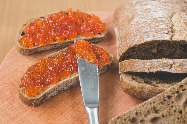 Red caviar toasts sandwich with silver knife, bowl of tasty red caviar with spoon and fresh baked homemade healthy bread on a wooden table background — Stock Photo, Image