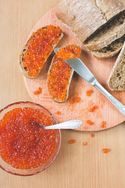 Красная икра тосты сэндвич с серебряным ножом, миска вкусной красной икры с ложкой и свежей выпечки домашнего здорового хлеба на деревянном столе фоне — стоковое фото