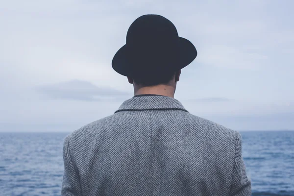 Stilig ung hipster guy i hatt tittar på disiga solsken genom en tjock dimma på ett lugnt hav och blå himmel bakifrån — Stockfoto