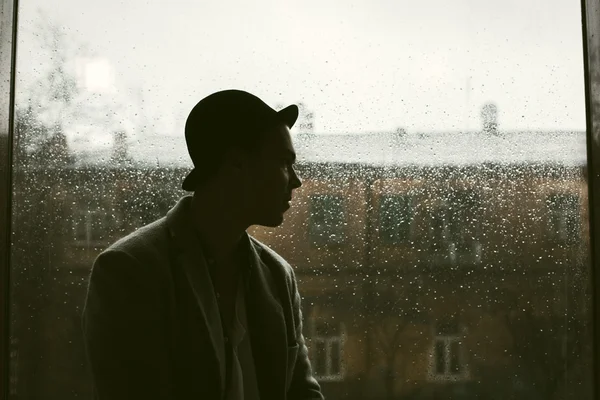 Retrato de um cara bonito caucasiano em silhueta chapéu em uma chuva cai na janela e cidade vista de fundo — Fotografia de Stock