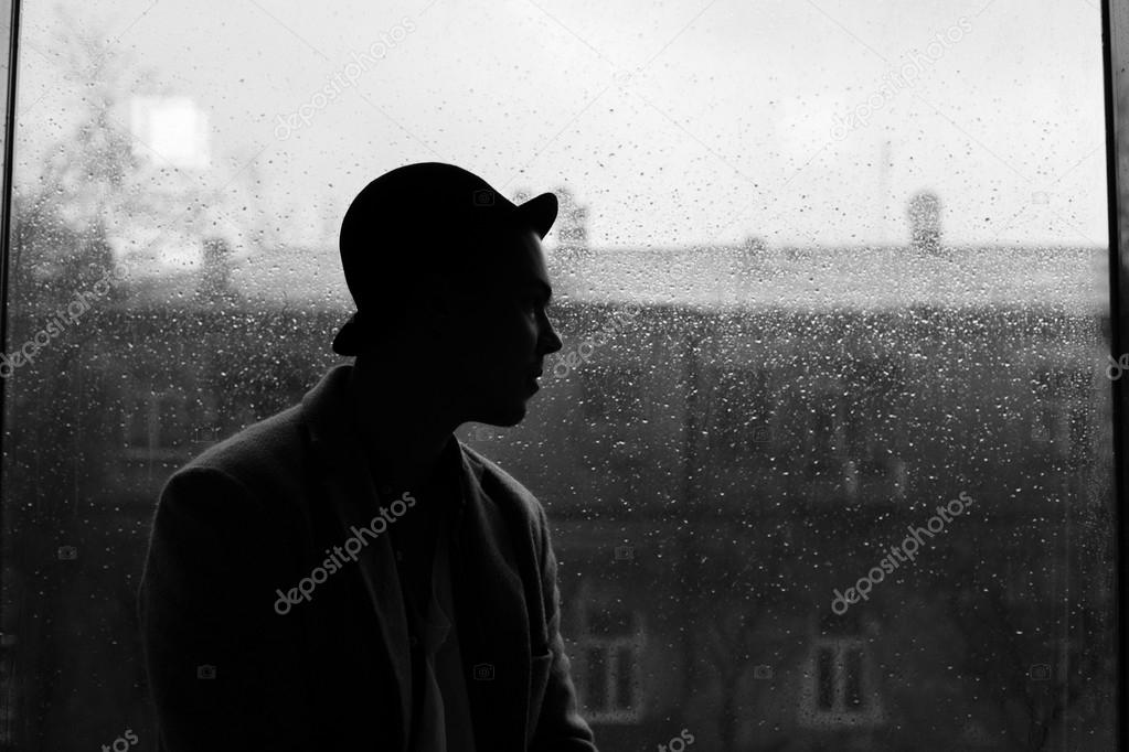 portrait of a handsome caucasian guy in hat silhouette on a rain drops on window and city view background