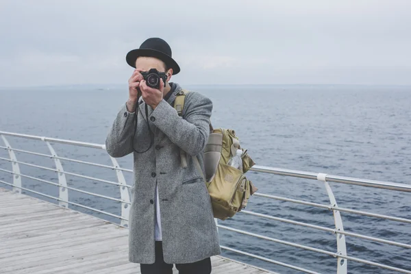 Handsome young hipster guy in hat taking picture with film camera of the sea — Stock Photo, Image