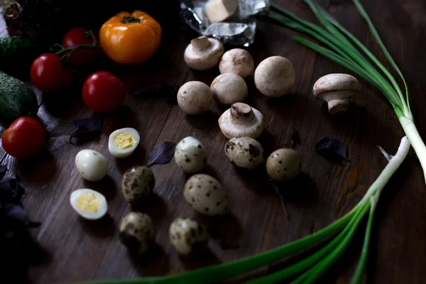 Preparación de ensaladas con huevos de codorniz, champiñones, mezcla de tomates rojos amarillos y cereza, pepino, albahaca, queso brie, lechuga y aceitunas negras, todos orgánicos y frescos del mercado de agricultores en rústico sryle sobre fondo de madera oscura enfoque suave en ángulo superior — Foto de Stock