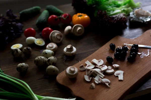 Preparación de ensaladas con huevos de codorniz, champiñones, mezcla de tomates rojos amarillos y cereza, pepino, albahaca, queso brie, lechuga y aceitunas negras, todos orgánicos y frescos del mercado de agricultores en rústico sryle sobre fondo de madera oscura enfoque suave en ángulo superior — Foto de Stock