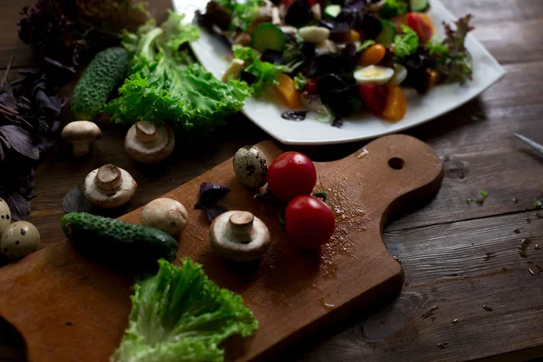 Preparación de ensaladas con huevos de codorniz, champiñones, mezcla de tomates rojos amarillos y cereza, pepino, albahaca, queso brie, lechuga y aceitunas negras, todos orgánicos y frescos del mercado de agricultores en rústico sryle sobre fondo de madera oscura enfoque suave en ángulo superior — Foto de Stock