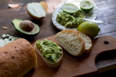 hazırlanması sürecinde avokado guacamole tablo kireç, soğan, avokado tüm organik ve taze koyu ahşap rustik tarzı arka plan üzerinde bir taze pişmiş ciabatta üzerinde hazır guacamole ile tüm ingridients yumuşak odak ek yük-açısı shot ekmek.