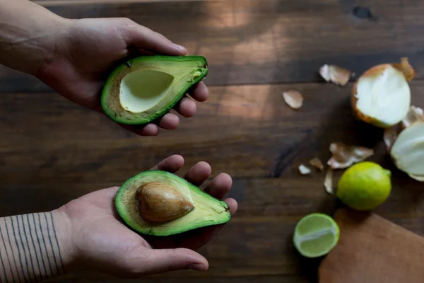 Preparación de aguacate guacamole en proceso todos los ingridientes sobre lima de mesa, cebolla, aguacate todo orgánico y fresco sobre fondo rústico de madera oscura con manos masculinas sosteniendo aguacate enfoque suave tiro de ángulo superior —  Fotos de Stock