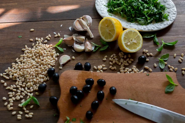 Italian pesto preparation vegan version without parmesan cheese with black olives instead all ingredients on table green basil, garlic, lemon, pine nuts, black olives all fresh and organic on dark wood rustic style table background soft focus — Stock Photo, Image